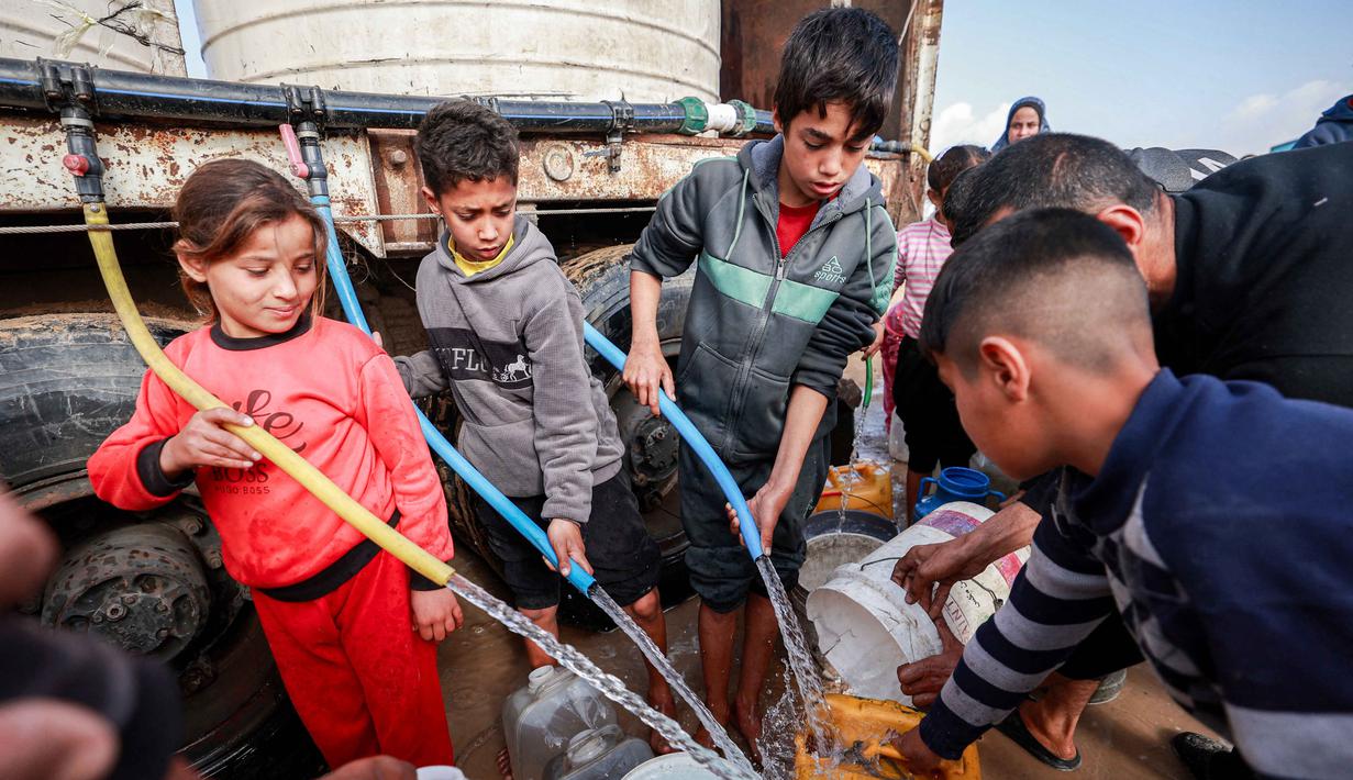 Sementara, kantor PBB untuk Koordinasi Urusan Kemanusiaan (OCHA) mengatakan bahwa para mitranya di sektor air, kebersihan, dan sanitasi melaporkan masih terjadinya kekurangan air bersih untuk kebutuhan rumah tangga, bahkan setelah jalur pasokan Mekorot dari Israel dibuka kembali. Tampak dalam foto, anak-anak menggunakan selang untuk mengisi wadah air dari tangki air portabel di kamp pengungsi Palestina Maghazi, Jalur Gaza tengah pada Rabu 11 Februari 2026. (Eyad BABA/AFP)