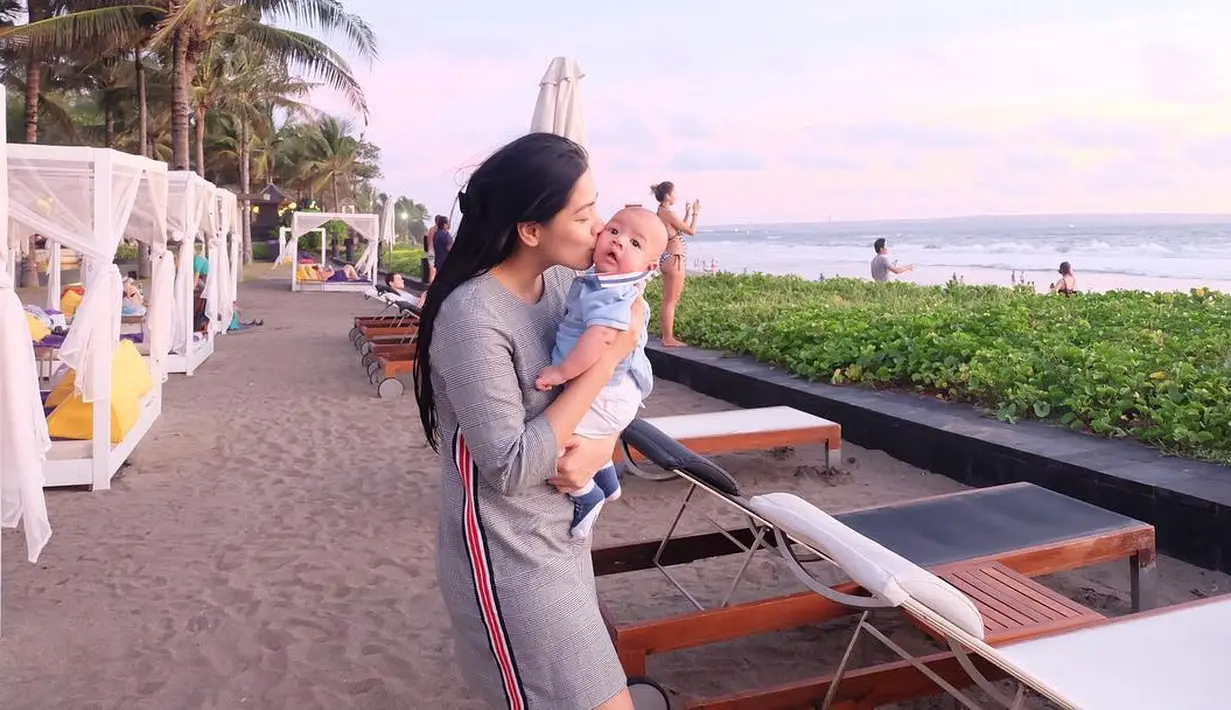 Nah kalau yang satu ini, terlihat Titi sedang menggendong buah hatinya sedang berlibur. Titi tetap cantik dengan dress abu-abunya saat menggendong Kai di tepi pantai. Lucu banget ya? (Instagram/titi_kamall)