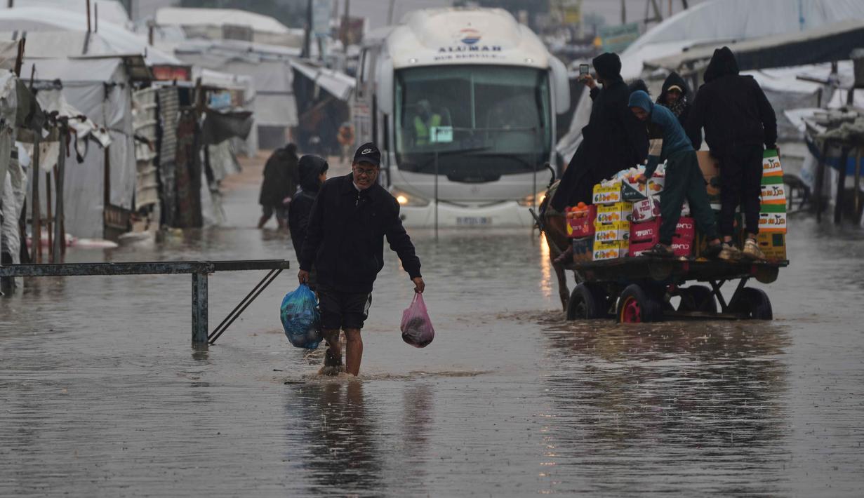 Situasi ini memperburuk genangan air di dalam dan sekitar tenda. Tampak dalam foto, warga Palestina berjalan melintasi jalan yang tergenang banjir setelah hujan lebat di Khan Younis, Jalur Gaza selatan, Kamis 11 Desember 2025. (AP Photo/Abdel Kareem Hana)