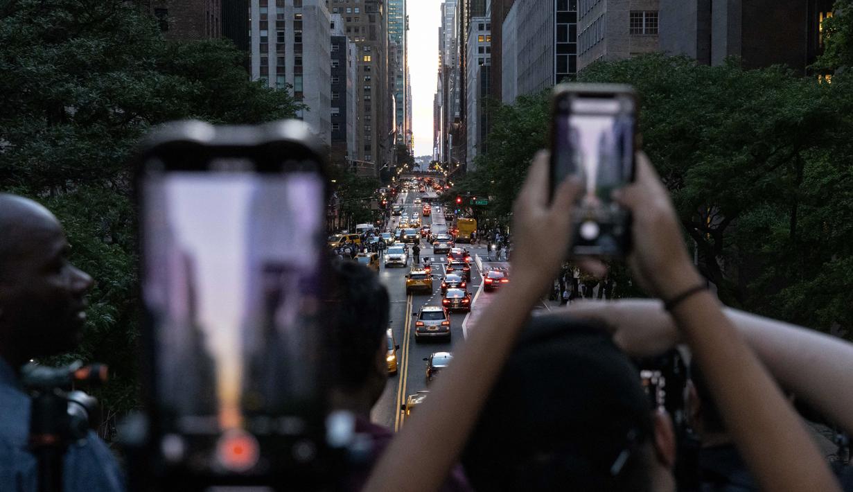 Orang-orang mengambil gambar saat matahari terbenam di Manhattan ketika fenomena "Manhattanhenge" di 42nd street, New York, Senin (11/7/2022). Fenomena Manhattanhenge hanyaterjadi di musim panas, tepatnya diantara bulan Mei hingga Juli. (Yuki IWAMURA / AFP)