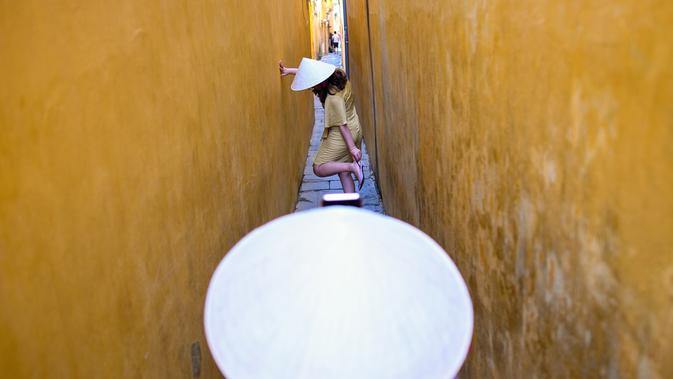 Seorang wanita mengenakan topi tradisional 'Non La' sambil berpose di lorong yang dicat warna kuning di kota tua Hoi An, 14 September 2019. Kota Tua Hoi An merupakan destinasi wisata populer di Vietnam yang mendapat gelar sebagai salah satu situs warisan dunia dari UNESCO. (Manan VATSYAYANA/AFP)