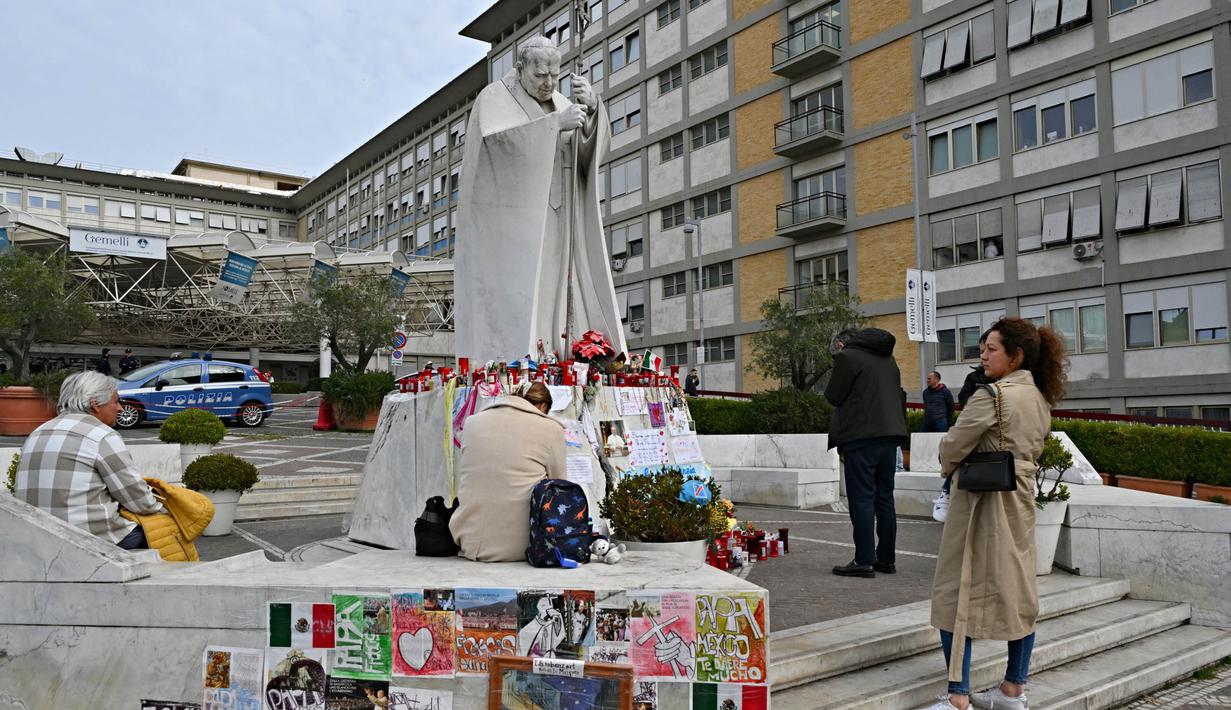 Orang-orang berdiri di depan patung Yohanes Paulus II di luar Rumah Sakit Universitas Gemelli tempat Paus Fransiskus dirawat karena pneumonia, di Roma pada 8 Maret 2025. (ANDREAS SOLARO/AFP atau pemberi lisensi/AFP)