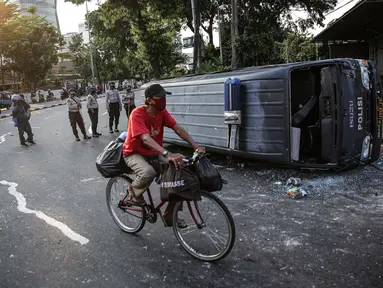 Seorang pengendara sepeda melintas dekat sebuah mobil polisi yang dirusak massa saat bentrok di kawasan Pejompongan, Jakarta, Rabu (7/10/2020). (Liputan6.com/Faizal Fanani)
