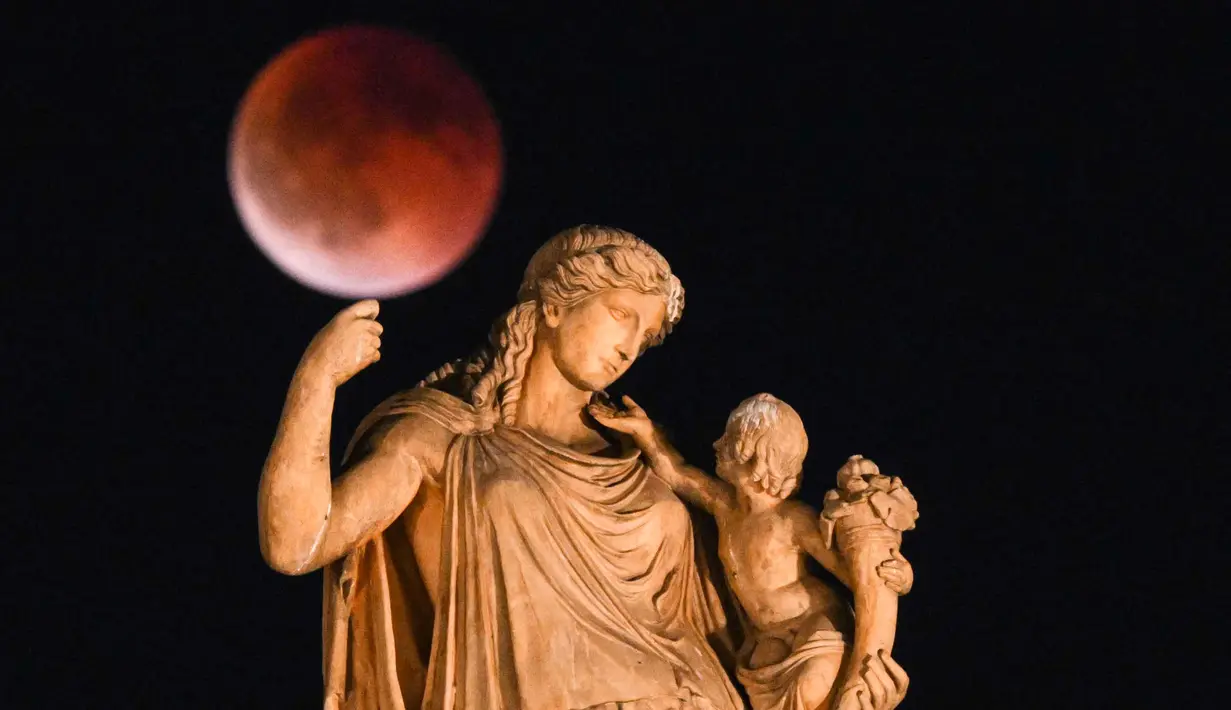 Foto ini menampilkan bulan purnama yang juga dikenal sebagai “Blood Moon” selama gerhana bulan, dengan patung dewi Yunani kuno Irene yang memegang seorang anak di latar depan, di pusat Athena, Yunani pada 7 September 2025. (Angelos TZORTZINIS/AFP)
