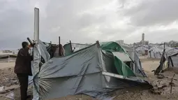 Ribuan tenda yang menjadi tempat tinggal keluarga pengungsi di Jalur Gaza rusak parah tersapu angin akibat badai musim dingin yang kuat. Tampak dalam foto, seorang pria Palestina yang mengungsi memperbaiki tenda yang didirikan di sepanjang pantai Kota Gaza saat angin musim dingin yang kencang menerpa wilayah tersebut pada Selasa 13 Januari 2026. (Omar AL-QATTAA/AFP)