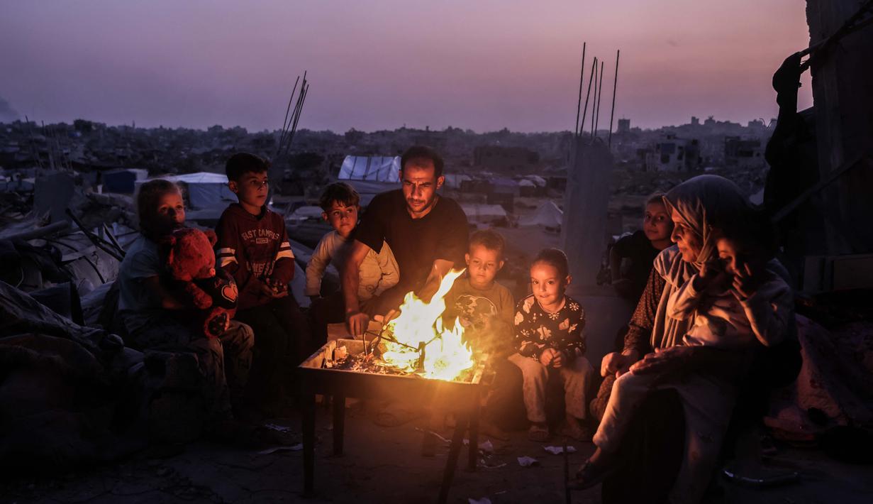 Farhan Haq juga menekankan bahwa PBB membutuhkan gencatan senjata berkelanjutan serta lebih banyak jalur penyeberangan yang terbuka agar rencana tanggap darurat 60 hari dapat dijalankan secara penuh. Tampak dalam foto, sebuah keluarga Palestina duduk di sekitar api unggun di tengah puing-puing bangunan yang hancur di Jabalia, di bagian utara Jalur Gaza, pada Jumat 7 November 2025. (Omar AL-QATTAA/AFP)
