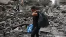 Seorang warga Palestina berjalan dengan membawa kantong plastik berisi barang-barang di tengah puing-puing bangunan yang hancur akibat serangan udara Israel di kawasan al-Rimal, Kota Gaza, pada 10 Oktober 2023. (MOHAMMED ABED/AFP)