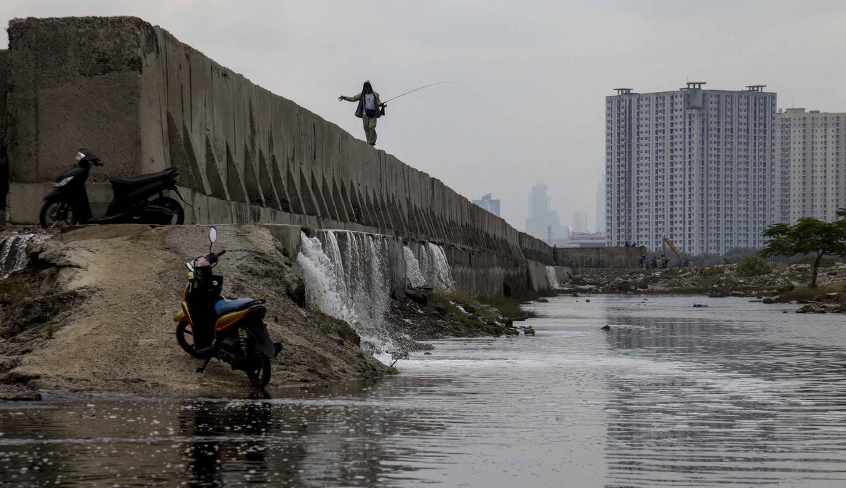 Namun, kali ini, Banjir rob di kawasan Muara Baru Jakarta diperparah dengan kebocoran dan rembesan pada tanggul laut di beberapa titik di Muara Baru, yang disebabkan oleh usia tanggul yang sudah tua dan korosi. Tampak dalam foto, seorang pria dengan pancingnya berjalan di atas tanggul laut raksasa yang dibangun untuk mencegah banjir di pesisir utara, seperti terlihat di Muara Baru, Jakarta pada Sabtu 6 Desember 2025. (ADITYA IRAWAN/AFP)