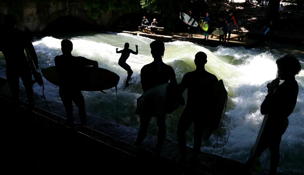 Sejumlah peselancar melihat rekannya menaklukkan ombak buatan saat bermain surfing di sungai 'Eisbach' di taman English Garden di Munich, Jerman (24/6/2019). Sungai Eisbach dipakai sebagai tempat berselancar sejak 1972 namun baru secara resmi diperbolehkan pada 2010. (AP Photo/Matthias Schrader)