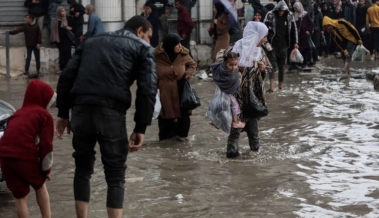 Ribuan warga pengungsi berjuang menahan masuknya air ke dalam tenda-tenda darurat di tengah kondisi kehidupan yang semakin berat menjelang musim dingin. Tampak dalam foto, orang-orang berjalan di tengah banjir di kamp darurat yang menampung warga Palestina yang terlantar setelah hujan deras di Kota Gaza pada Selasa 25 November 2025. (Omar AL-QATTAA/AFP)