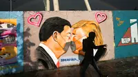 Seorang pria berjalan di depan Graffiti yang menggambarkan Presiden AS Trump (kanan) dan Presiden Tiongkok Xi Jinping saling berciuman dengan mengenakan masker di tembok taman umum Mauerpark di Berlin, Jerman, Rabu, (29/4/2020). (AP/Markus Schreiber)