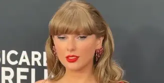 Taylor Swift hadir mengenakan mini dress merah yang berkilauan model one shoulder dari Vivienne Westwood. Ia melengkapi tampilannya dengan aksesori anting-anting merah dan aksesori di bagian kakinya yang mencuri perhatian. Aksesori tersebut memiliki liontin huruf T sesuai namanya dan nama kekasihnya. [@voguemagazine]