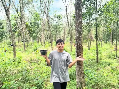 Dalam salah satu video, Jirayut juga membagikan suasana di area kebun. Jirayut berdiri di kebun karetnya yang luas. Latar belakang memperlihatkan puluhan pohon karet yang berjajar rapi. Langit yang cerah dan rumput hijau menambah kesan natural dan tenang. (Liputan6.com/IG/@jirayutdaa4official)
