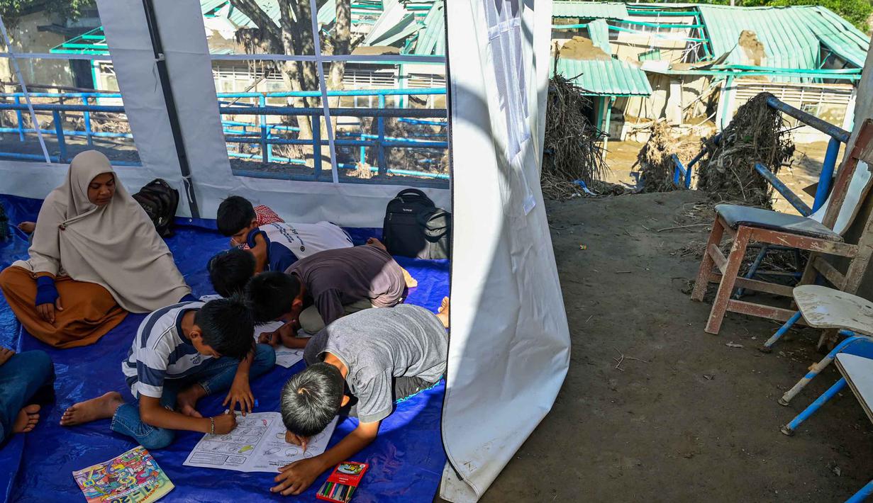 Kegiatan belajar mengajar terpaksa dilakukan di tenda-tenda darurat. Tampak dalam foto, para siswa berkumpul di kelas darurat di dalam tempat penampungan setelah sekolah mereka hancur akibat banjir bandang di daerah yang berdekatan dengan Sungai Peusangan di Bireuen, provinsi Aceh, Senin 5 Januari 2026. (CHAIDEER MAHYUDDIN/AFP)
