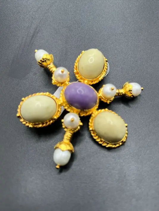 Bros mewah berbentuk bunga dengan perpaduan batu alam Kalimantan berwarna krem dan ungu di tengah, dilengkapi mutiara imitasi yang memancarkan kesan anggun. Bingkai berlapis emas menambah kilau elegan, menjadikannya aksesori sempurna untuk melengkapi busana pesta maupun acara spesial. [Dok/batualamkalimantan]