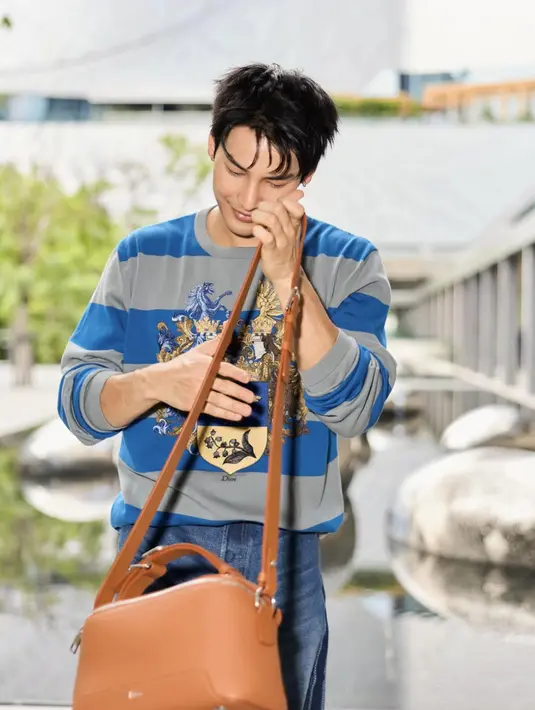 Aktor Thailand, Apo Nattawin tampil dengan atasan lengan panjang bergaris biru abu-abu dipadukan celana denim, sambil membawa Dior Archie Bag coklat. [@apo555]