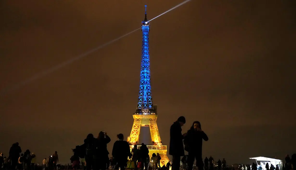 Para turis berada di sekitar Menara Eiffel diterangi dengan warna-warna khas Ukraina untuk menandai peringatan satu tahun invasi Rusia ke negara tersebut, di Paris, Kamis, 23 Januari 2023. Pada 24 Februari 2022 lalu Rusia menginvasi Ukraina dan menandai puncak eskalasi konflik kedua negara sejak 2014 silam. (AP Photo/Christophe Ena)