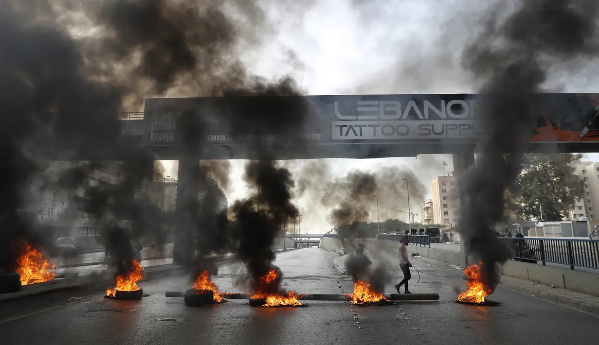 Pengunjuk rasa lewat depan ban yang terbakar di jalan raya utama yang mengarah ke Bandara Internasional Beirut selama protes terhadap kenaikan harga barang-barang konsumsi dan jatuhnya mata uang lokal di Beirut, Lebanon, Senin (29/11/2021). (AP Photo/Hussein Malla)