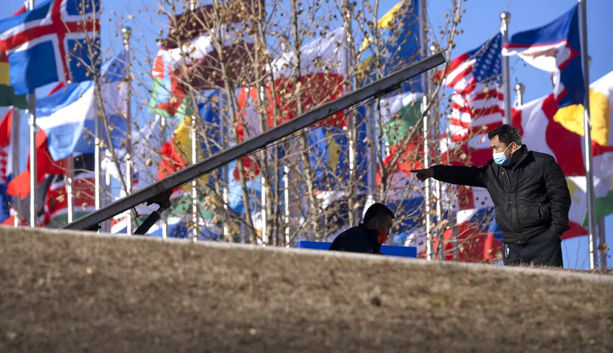 Para pekerja yang mengenakan masker berbicara dekat pajangan bendera negara-negara peserta di alun-alun terbuka di Desa Olimpiade Musim Dingin, Beijing, China, Jumat, 24 Desember 2021. Olimpiade dan Paralimpiade Musim Dingin 2022 akan diadakan mulai bulan Februari. (AP Photo/Mark Schiefelbein)