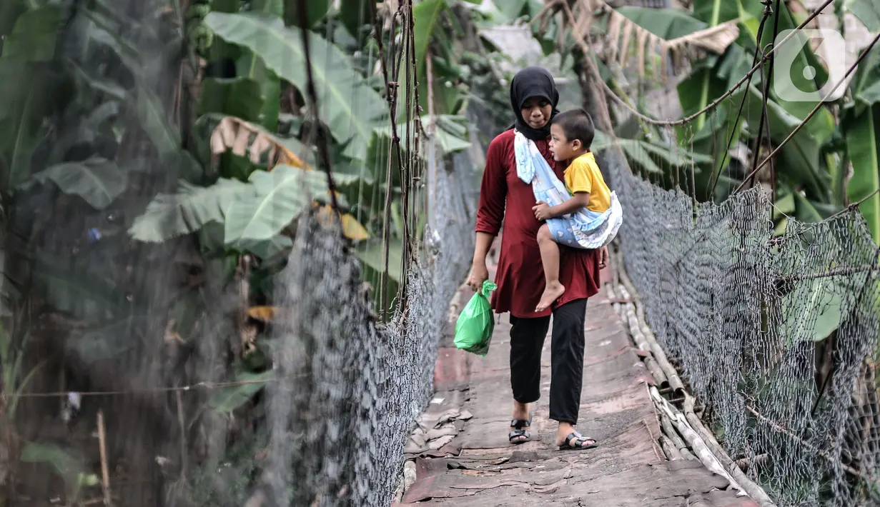 Warga menggendong anaknya saat melintasi jembatan gantung yang sudah tidak layak di kawasan Srengseng Sawah, Jakarta, Sabtu (12/6/2021). Kondisi jembatan gantung di atas Sungai Ciliwung itu saat ini tidak layak dan dapat membahayakan keselamatan warga yang melintas. (merdeka.com/Iqbal S. Nugroho)