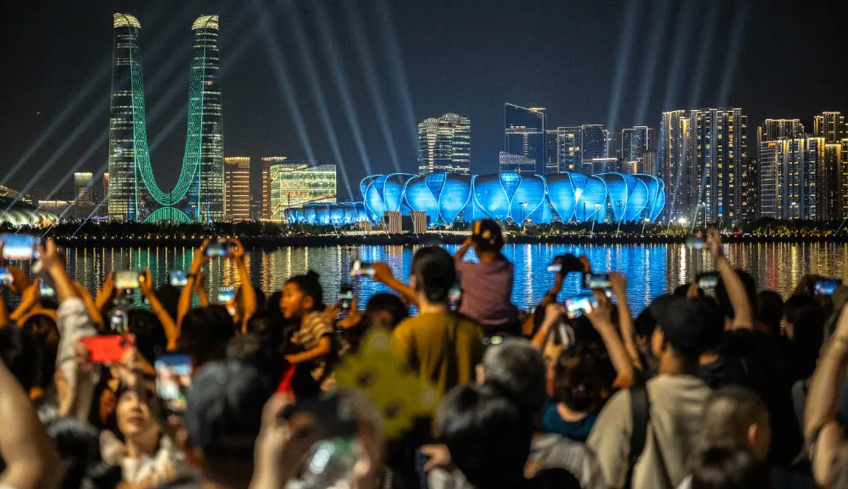 Orang-orang berkumpul di kawasan pejalan kaki Sungai Qiantang untuk menyaksikan pertunjukan cahaya di Hangzhou Olympic Sports Centre Stadium (kanan) menjelang Asian Games 2023 di Hangzhou, Provinsi Zhejiang, China, Selasa (19/9/2023). Hangzhou Olympic Sports Centre Stadium merupakan lokasi pembukaan Asian Games 2023. (Philip FONG/AFP)