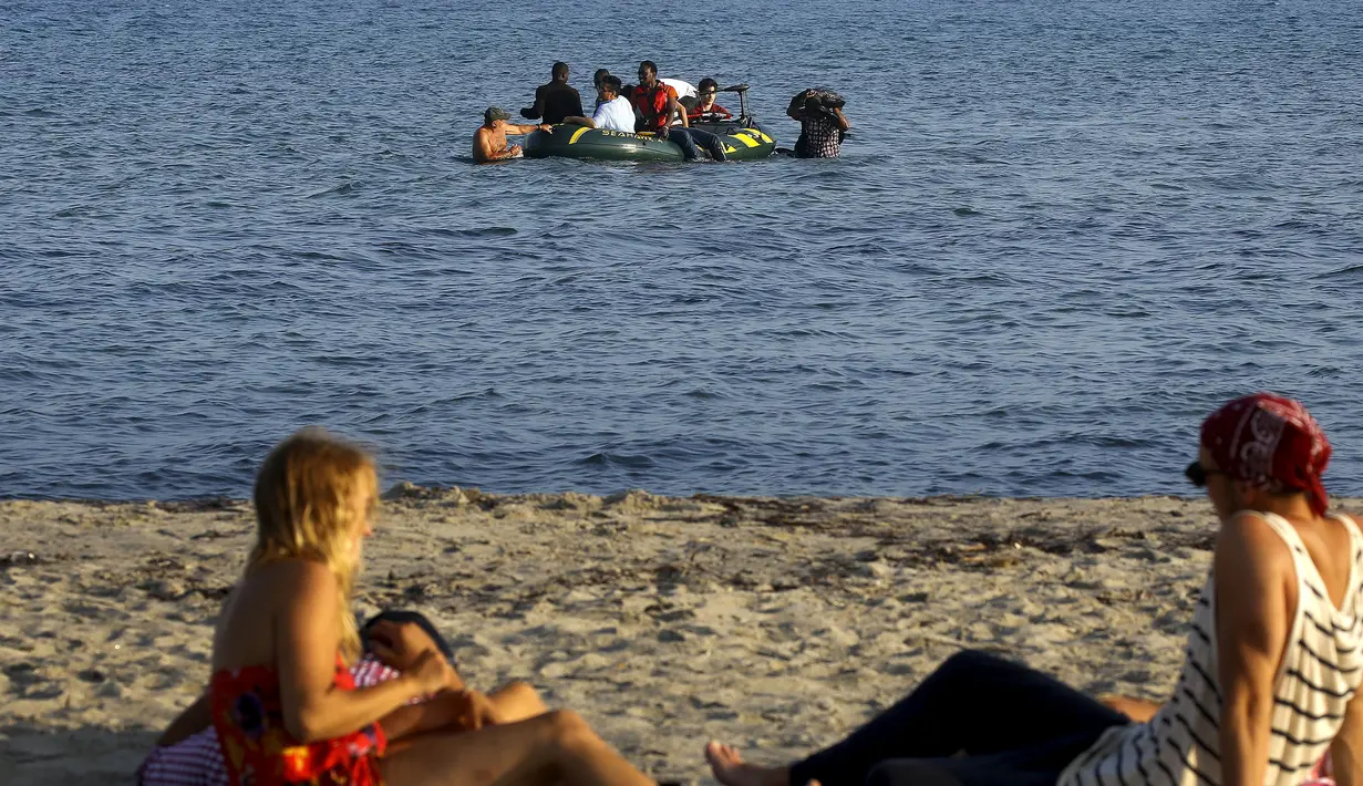 Wisatawan berbaring di pantai melihat para imigran dan pengungsi dari Suriah dan Afrika tiba saat berada diperahu usai melintasi Laut Aegea antara Turki dan Yunani, (8/8/2015). (REUTERS/Yannis Behrakis)