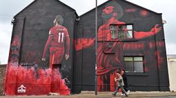 Dua pejalan kaki melewati mural seniman Inggris, John Culshaw yang menggambarkan pesepak bola Mesir Mohamed Salah di fasad bangunan dekat Stadion Anfield, di Liverpool, Rabu (30/3/2022). Salah sudah berada di urutan kesembilan dalam daftar pencetak gol sepanjang masa Liverpool. (Paul ELLIS / AFP)