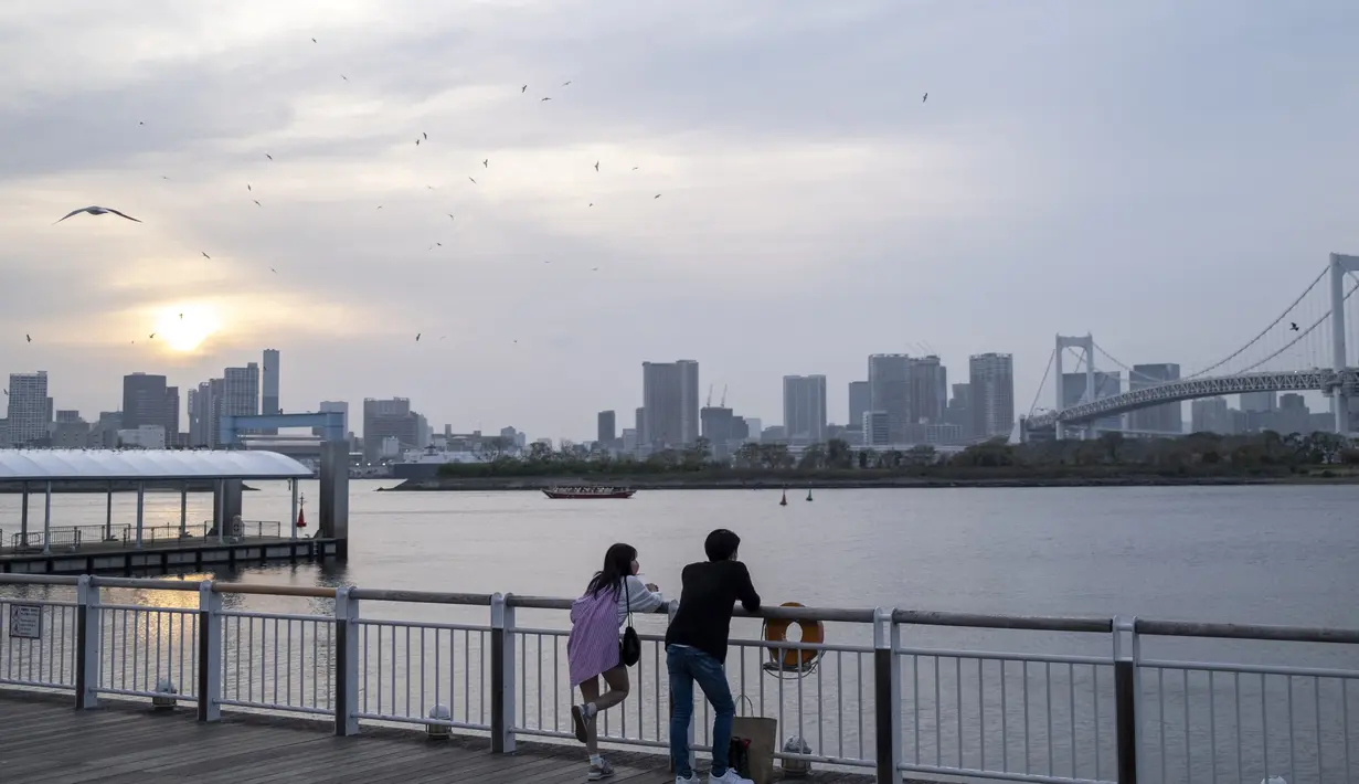 Orang-orang melihat matahari terbenam di dekat jembatan Pelangi di daerah Odaiba Tokyo (11/4/2022). Odaiba yang dapat diakses dari Jembatan Pelangi atau kereta Yurikamome yang futuristik adalah pusat hiburan teknologi canggih di sebuah pulau buatan di Teluk Tokyo. (AFP/Charly Triballeau)