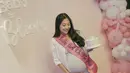 Saat baby shower, sambil memegang cake pink, Winona Willy mengenakan pakaian serba putih dari kemeja lengan pendek dipadukan inner dress panjang serta flat shoes ber jaringnya. [@nonawilly]