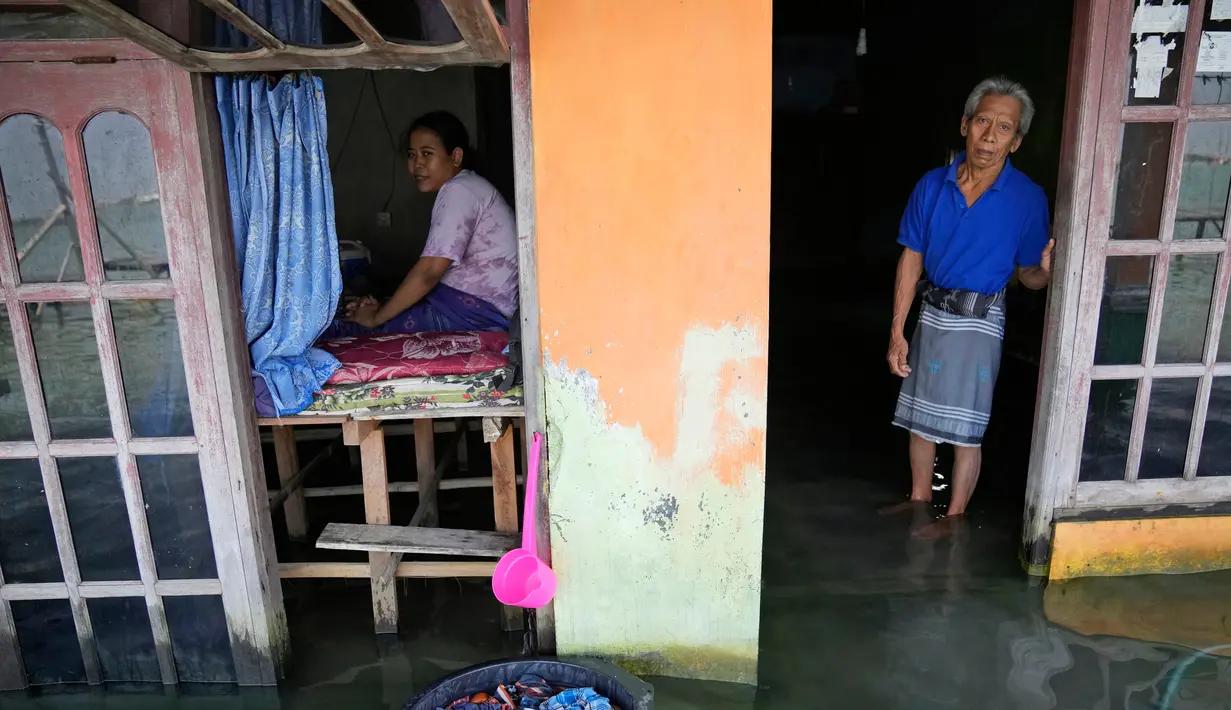 Sudarto berdiri di depan pintu rumahnya yang terendam banjir saat putrinya Turiah memandangi rumah mereka yang terendam banjir di Timbulsloko, Jawa Tengah, Indonesia, 31 Juli 2022. Dengan keterbatasan fisik yang menghalangi Turiah dari pekerjaan normal di desa, ia menghabiskan harinya dengan duduk di jendela depan rumah di atas panggung kayu yang ditinggikan. (AP Photo/Dita Alangkara)