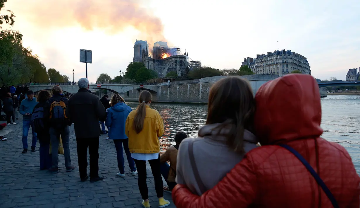 Orang-orang menyaksikan api dan asap mengepul dari kebakaran Gereja Katedral Notre-Dame di pusat kota Paris, Prancis, pada Senin (15/4). Api yang melalap bagian atas gereja kuno itu membuat kerumunan baik warga Paris maupun turis tertegun. (AP Photo/Thibault Camus)