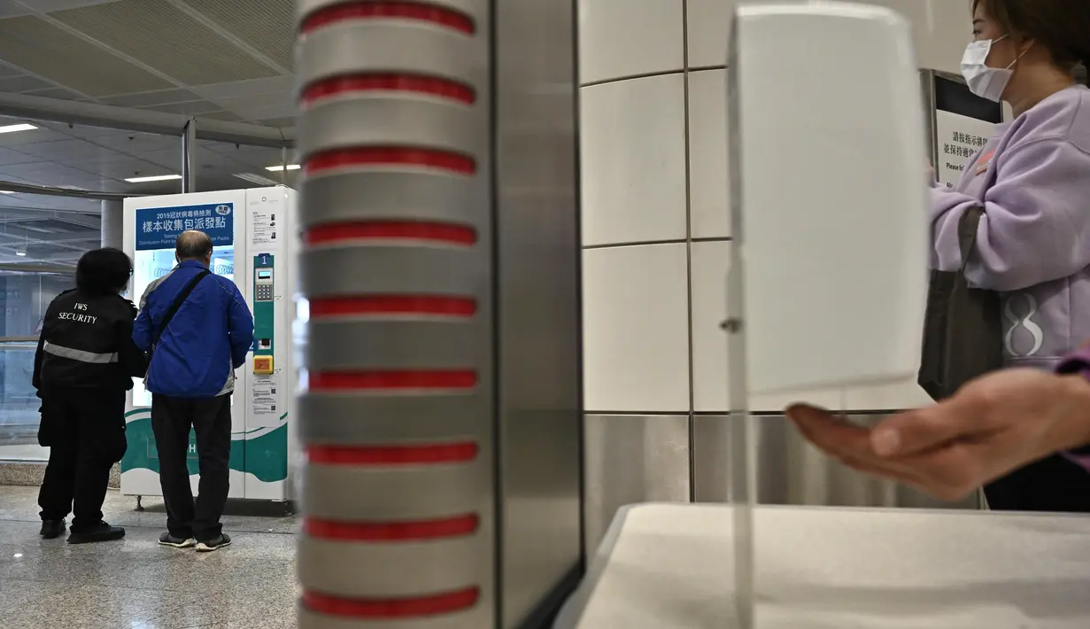 Seseorang (kanan) memakai pembersih tangan sambil menunggu untuk menggunakan mesin penjual otomatis (kiri) yang menyediakan alat test kit COVID-19 di stasiun kereta bawah tanah MTR di Hong Kong pada Senin (7/12/2020). (Anthony WALLACE / AFP)