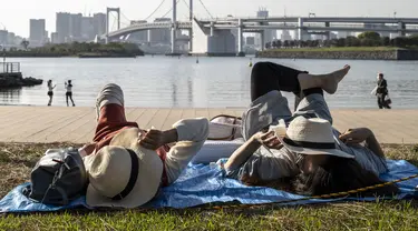 Orang-orang berbaring di rumput pada hari yang cerah di tepi teluk di daerah Odaiba Tokyo (12/4/2022). Odaiba yang dapat diakses dari Jembatan Pelangi atau kereta Yurikamome yang futuristik adalah pusat hiburan teknologi canggih di sebuah pulau buatan di Teluk Tokyo. (AFP/Charly Triballeau)