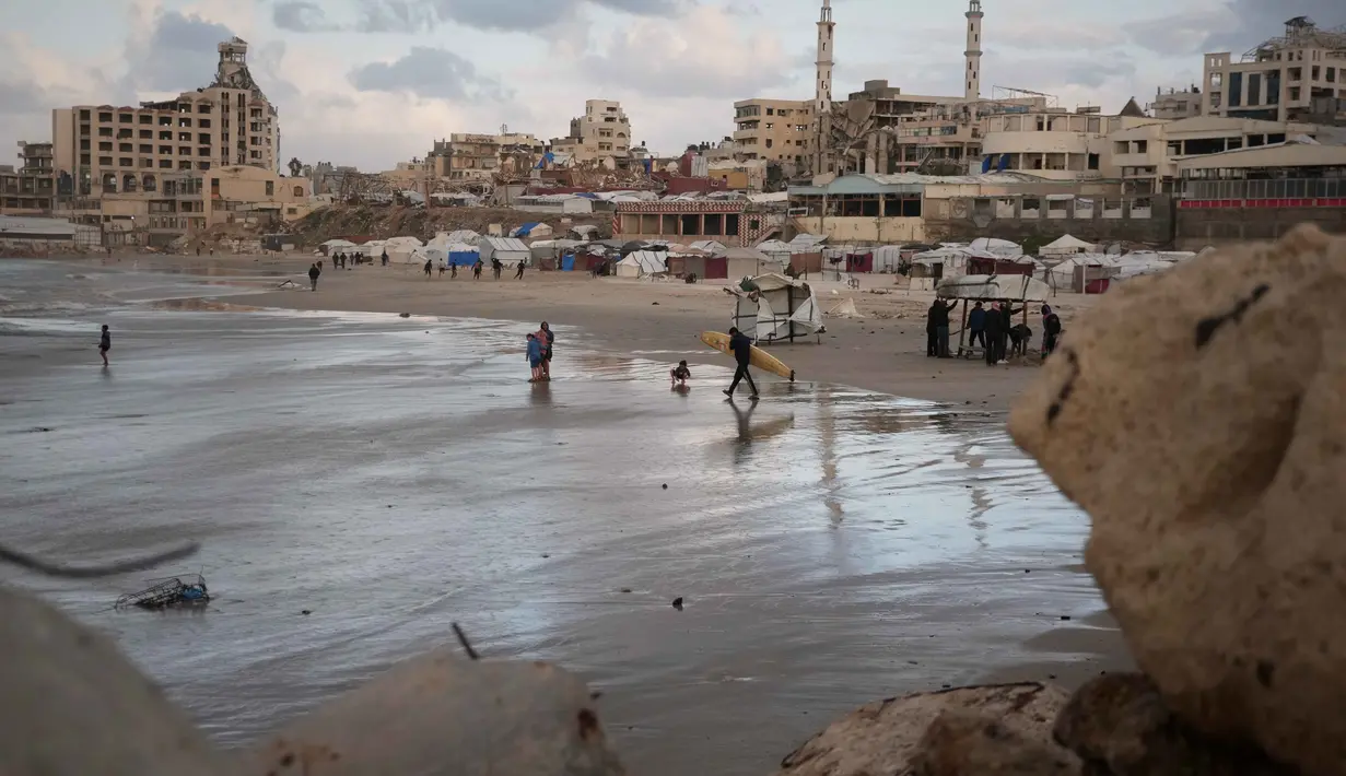 Warga Palestina menata kembali kehidupan mereka di antara puing-puing kehancuran akibat serangan yang dilancarkan Israel sejak Okrober 2023 silam. Tampak dalam foto, seorang peselancar Palestina membawa papan selancarnya ke laut di sepanjang garis pantai di Kota Gaza, Minggu, 28 Desember 2025. (AP Photo/Jehad Alshrafi)