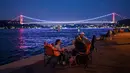 Orang-orang menikmati malam di sebelah pantai Bosphorus ketika jembatan Fatih Sultan Mehmet terlihat di Istanbul (6/7/2020). Jembatan Fatih Sultan Mehmet, yang dikenal dengan sebutan Jembatan Bosporus Kedua, adalah sebuah jembatan di Istanbul, Turki yang melintasi selat Bosporus. (AFP/Ozan Kose)