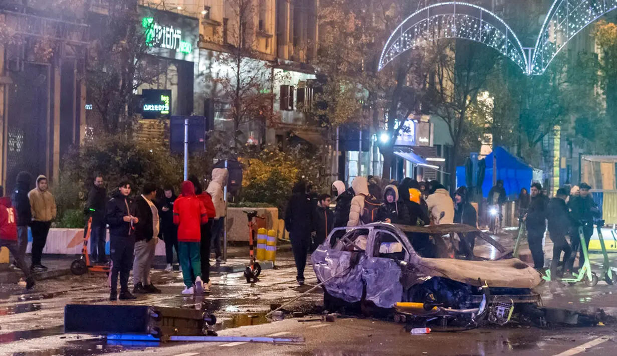 Orang-orang berdiri di samping mobil yang terbakar saat kerusuhan di Brussel, Belgia, 27 November 2022. Polisi harus menutup sebagian pusat Kota Brussel dan bergerak dengan meriam air serta gas air mata untuk membubarkan massa menyusul kerusuhan selama dan setelah Maroko menang 2-0 atas Belgia di Piala Dunia 2022. (AP Photo/Geert Vanden Wijngaert)