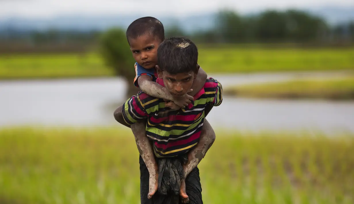 Seorang pemuda etnis Rohingya dari Myanmar menggendong anak kecil di punggungnya saat berjalan melewati sawah untuk menyebrang ke perbatasan Bangladesh, wilayah Teknaf yang berada di distrik Cox's Bazar, 1 September 2017. (AP Photo/Bernat Armangue)