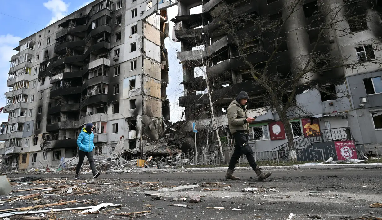 Orang-orang berjalan melewati bangunan yang hancur di kota Borodianka, barat laut Kiev pada 4 April 2022. Saat pasukan Rusia mundur, kota kecil Borodianka, 50 km barat laut Kiev, menjadi reruntuhan. (Sergei SUPINSKY / AFP)