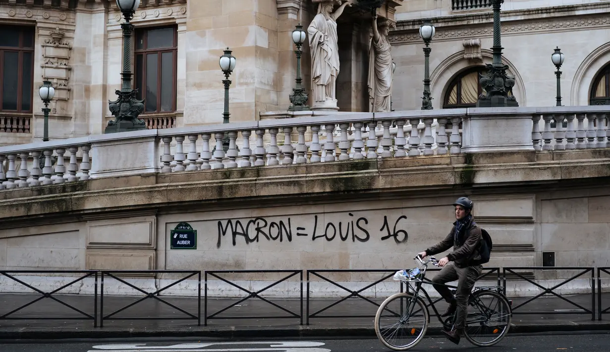 Seorang pengendara sepeda melewati grafiti bertuliskan 'Macron sama dengan Louis 16' di rumah Opera Garnier Paris, Prancis, Minggu (2/12). Hal itu merujuk pada Raja Prancis selama Revolusi Prancis tahun 1789. (AP Photo/Kamil Zihnioglu)