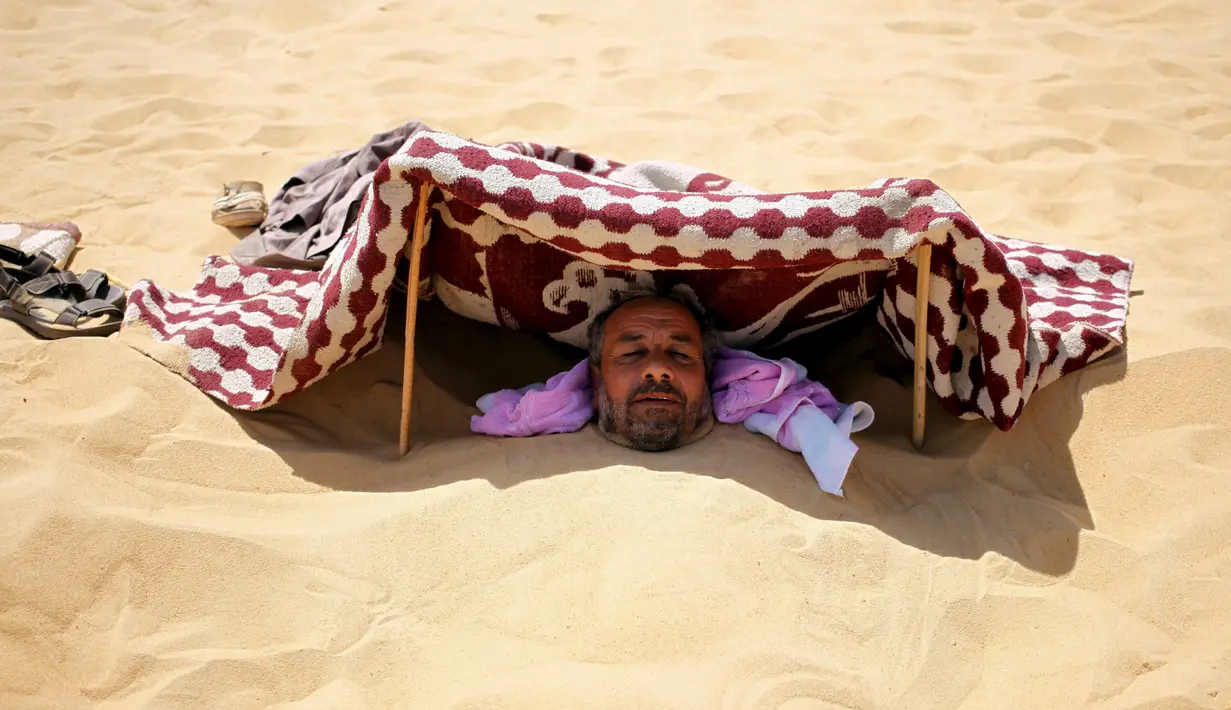 Seorang pria menutupi kepalanya saat dikubur dengan pasir gurun di Siwa, Mesir, 12 Agustus 2015. Terapi mengubur diri di dalam pasir gurun saat terik matahari diyakini dapat menyembuhkan rematik, sakit sendi, dan impotensi seksual. (REUTERS/Asmaa Waguih)