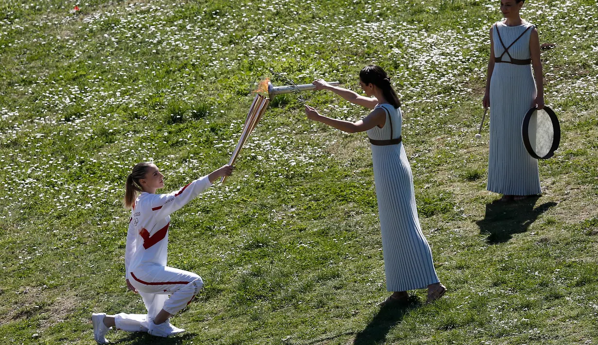 Aktris Yunani Xanthi Georgiou (tengah) sebagai Imam Besar menyalakan obor Olimpiade Tokyo 2020 yang dibawa juara menembak Olimpiade asal Yunani, Anna Korakaki (kiri) saat upacara penyalaan api Olimpiade Tokyo 2020 di Kota Tua Olympia, Yanani, Kamis (12/3/2020). (AP Photo/Yorgos Karahalis)