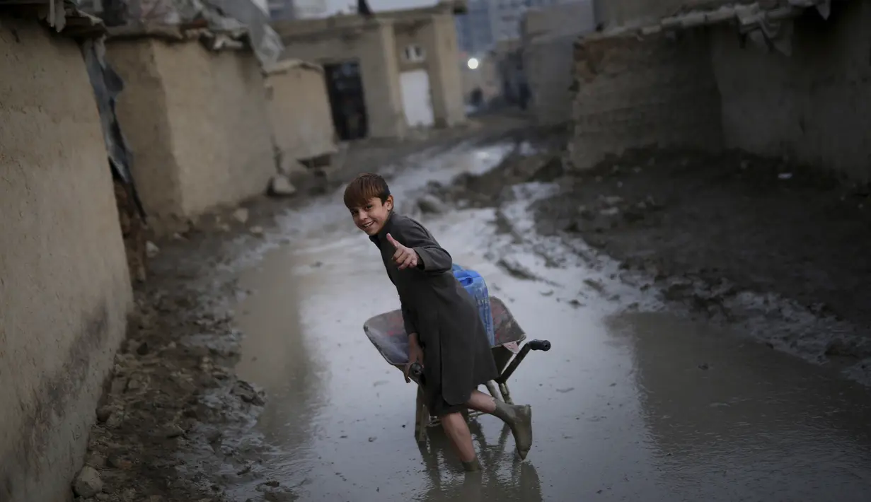 Seorang anak mengacungkan jempol saat mendorong gerobak melalui jalan berlumpur di sebuah kamp di Kabul, Afghanistan (27/11/2019). Puluhan ribu warga Afghanistan yang terlantar secara internal tinggal di kamp-kamp, yang kekurangan fasilitas dasar, di seluruh Afghanistan. (AP Photo/Altaf Qadri)
