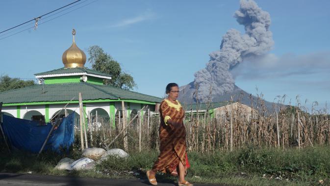 Seorang perempuan berjalan saat Gunung Sinabung mengeluarkan erupsi asap tebal ke udara di Karo, Sumatera Utara, Minggu, (23/8/2020). Untuk kesekian kalinnya Gunung Sinabung kembali erupsi dengan menyemburkan abu vulkanis ke udara hingga radius 2 kilometer. (Muhammad Zulfan Dalimunthe / AFP)