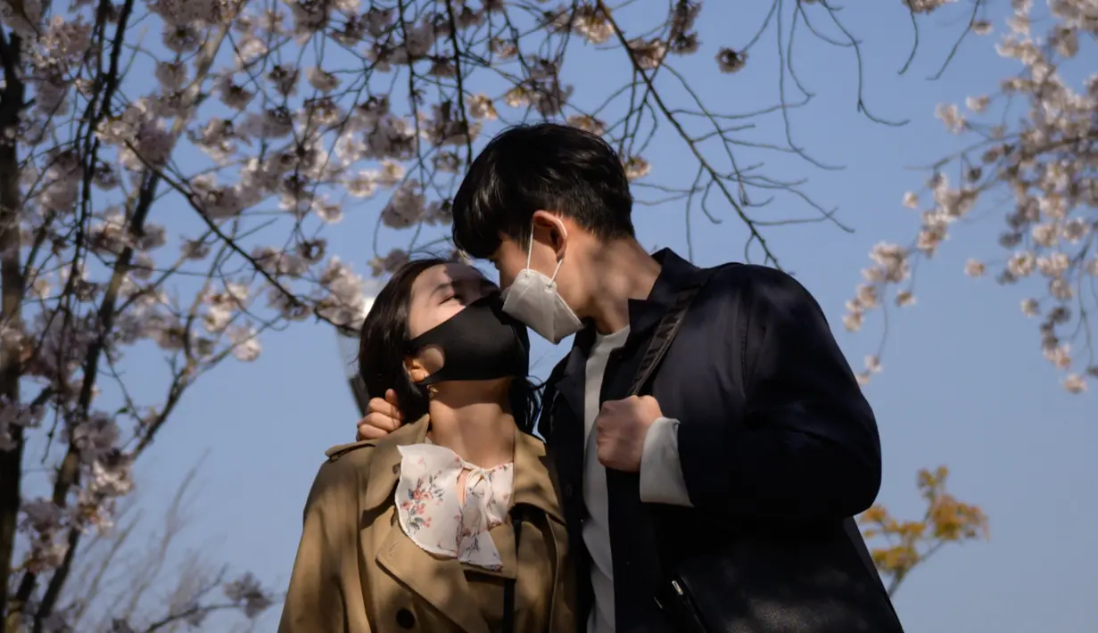 Pasangan mengenakan masker saat berdiri di bawah pohon yang mekar di Distrik Yeouido, Seoul, Korea Selatan, 5 April 2020. Organisasi Kesehatan Dunia (WHO) mengumumkan virus corona COVID-19 sebagai pandemi sejak 11 Maret 2020 lalu. (Ed JONES/AFP)