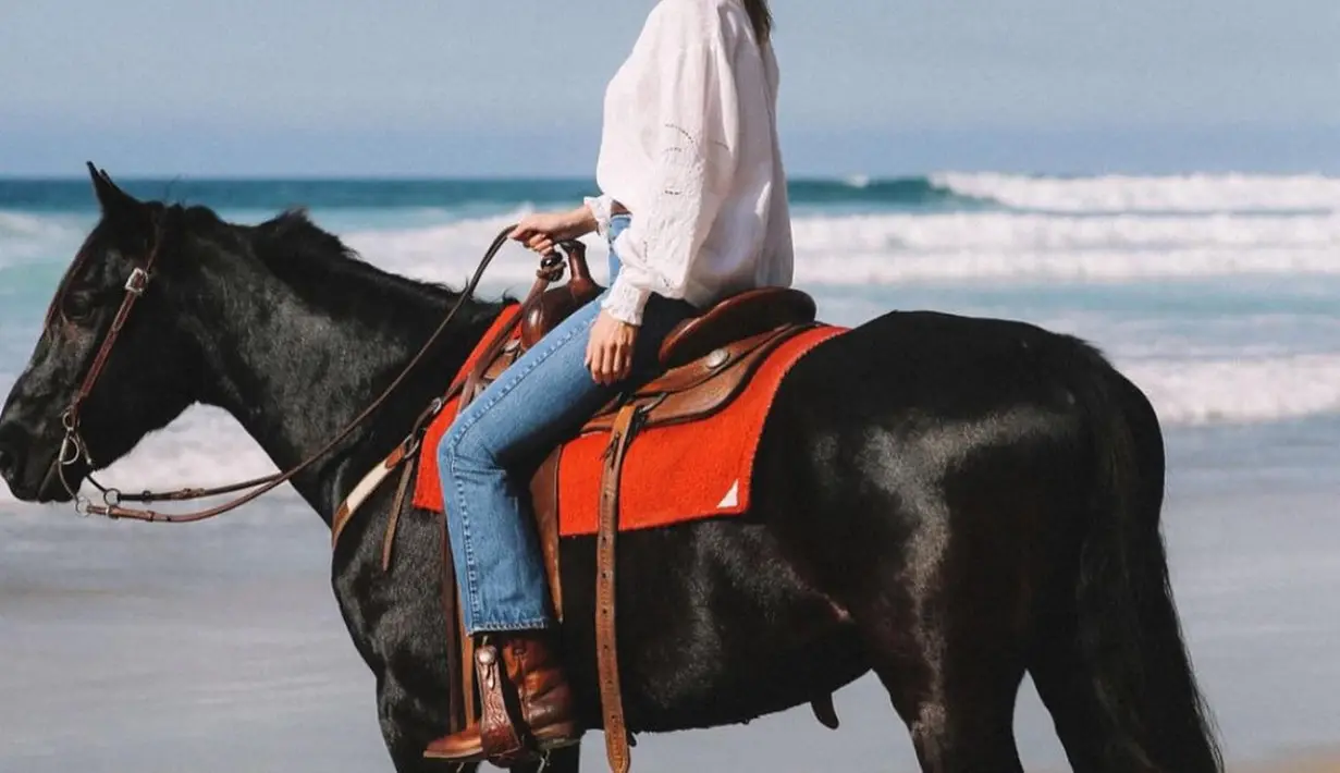 Alana Hadid dalam balutan atasan blouse putih dan celana jeans, dan midi boots cokelat, naik kuda hitam di pinggir pantai. [Foto: Instagram/lanzybear]