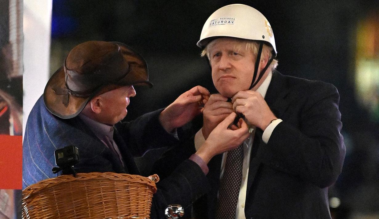 Perdana Menteri Inggris Boris Johnson (kanan) menerima bantuan saat ia mengenakan helm sebelum mengendarai sepeda saat kunjungannya ke pasar Makanan dan Minuman Inggris yang didirikan di Downing Street, London, 30 November 2021. Ada 12 bisnis dari seluruh Inggris. (JUSTIN TALLIS/AFP/POOL)