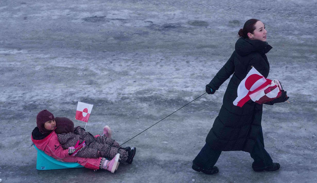 Donald Trump bahkan tidak menutup kemungkinan penggunaan kekuatan untuk menguasai wilayah tersebut. Tampak dalam foto, weorang wanita menarik anak-anaknya di atas kereta luncur saat demonstrasi menentang kebijakan Presiden Amerika Serikat (AS), Donald Trump, terhadap Greenland di depan konsulat AS di Nuuk, Sabtu 17 Januari 2026. (AP Photo/Evgeniy Maloletka)