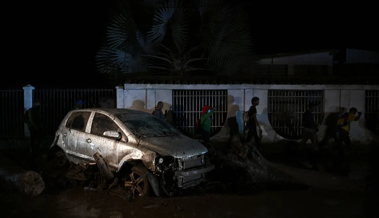 Orang-orang berjalan di samping mobil yang rusak oleh tanah longsor akibat hujan lebat di El Castaño, Venezuela, Senin (17/10/2022). Kini anggota tim penyelamat telah dikerahkan untuk memperluas batas pencarian mereka. (Federico Parra / AFP)