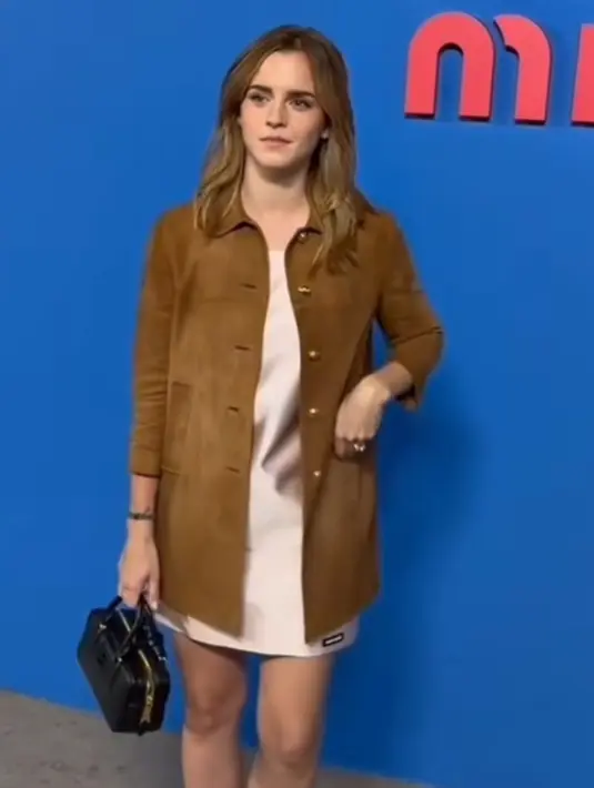 Emma Watson hadir mengenakan long outer coklat dipadukan inner mini dress soft pink. Ia membawa hand mini bag hitam senada dengan warna heels strapnya. [Dok. @wwd]