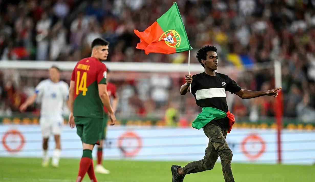 Seorang penyusup lapangan berlari sambil mengibarkan bendera Portugal di depan bek Portugal, Antonio Silva pada tengah laga Grup J Kualifikasi Euro 2024 antara Timnas Portugal menghadapi Bosnia Herzegovina di Luz Stadum, Lisbon, Portugal, Minggu (18/6/2023) dini hari WIB. (AFP/Patricia De Melo Moreira)
